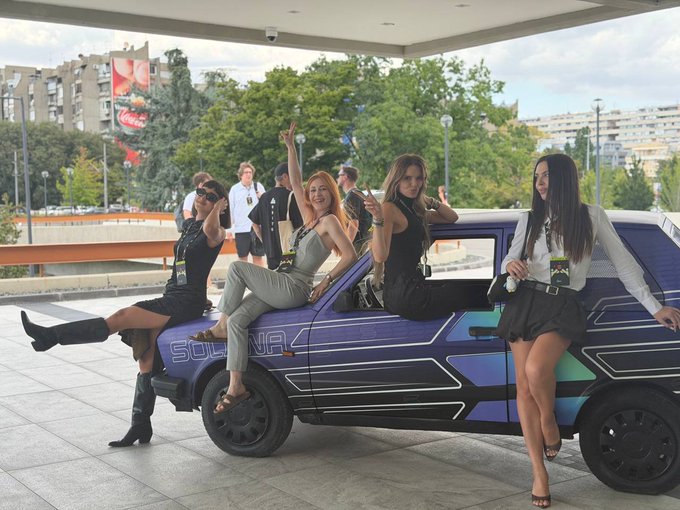 Four women pose on and around a purple car with 'SOLUNA' branding, with an urban background including trees and buildings.