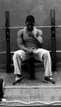 Black and white vintage photograph of a shirtless man in light-colored pants sitting on a wooden chair with his fist raised to his chin in a thinking pose.