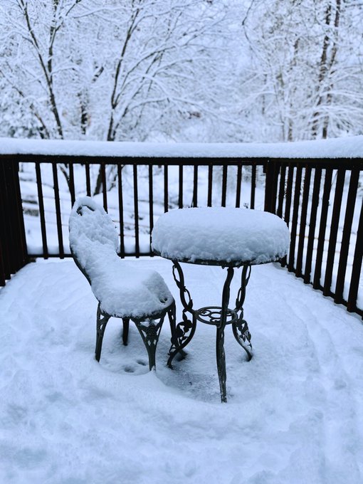 A snow-covered balcony scene showing a metal chair and small round table completely blanketed in snow, surrounded by snow-laden trees in the background.