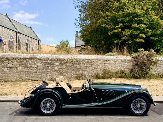 A dark green vintage Morgan-style roadster convertible parked on a street in front of a stone wall and an old stone church or chapel with a slate roof.