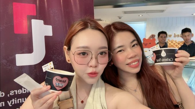 The image features two women posing for a picture with cups of ice cream, one holding a cup labeled "Just Us" while the other holds another cup that says "Baddies". The background includes a poster advertising a product or service related to blockchain technology.
