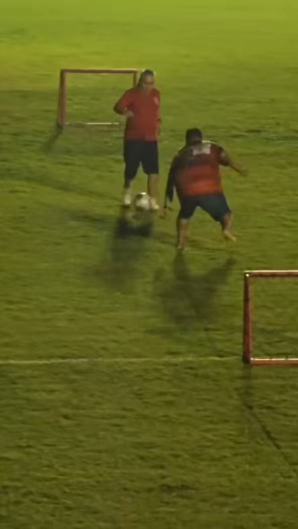 The image shows two men playing soccer on a field, with one man kicking a soccer ball towards another player who is attempting to block the shot. The scene takes place at night, adding an element of darkness to the atmosphere. In the background, there are red goal posts and a fence that separates the field from other areas in the vicinity. A caption below the image reads "Bitcoin bouncing between $60k and $80k".