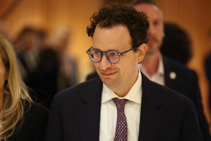 A man with curly hair and glasses wearing a dark suit and patterned tie is smiling slightly at an event.