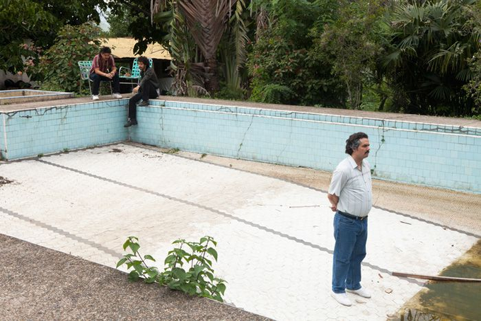 A photograph showing two men sitting by an empty, tiled swimming pool in the background, while a man in a white shirt and blue jeans stands looking at the empty pool in the foreground.