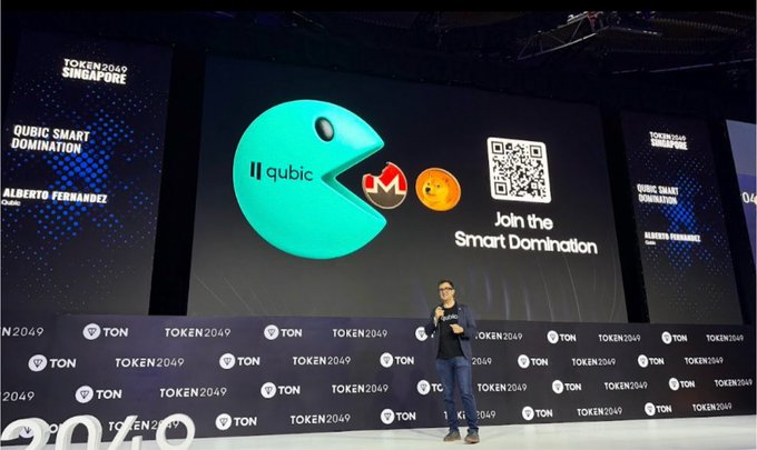 A speaker presents at TOKEN2049 Singapore, standing before a large screen displaying the "qubic" logo alongside a QR code and the slogan "Join the Smart Domination," tying the accompanying post's message about QUBIC driving next-gen decentralized AI to a live industry event.