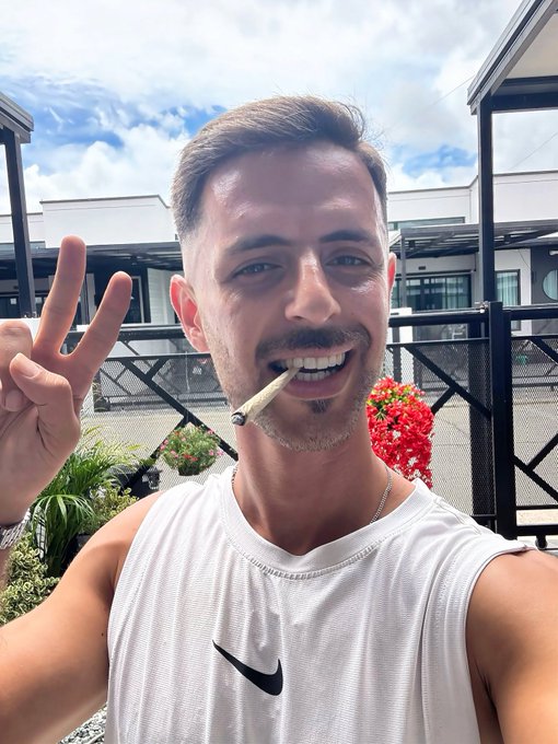 A selfie of a smiling young man with a mustache and goatee holding a peace sign, with a cigarette in his mouth, wearing a white Nike sleeveless shirt on a sunny rooftop.
