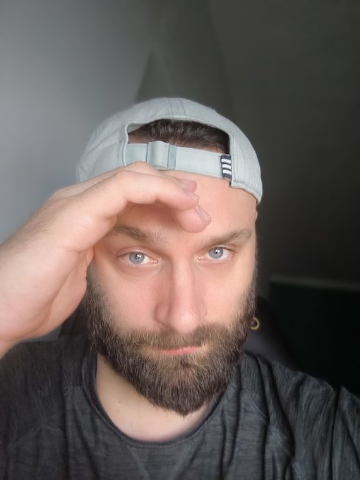 The image features a man with a beard wearing a white baseball cap that is slightly tilted towards his forehead as he holds up one of his hands, possibly touching or rubbing his face. The background appears to be a dark room, which contrasts with the bright subject matter in the foreground and brings attention to him.