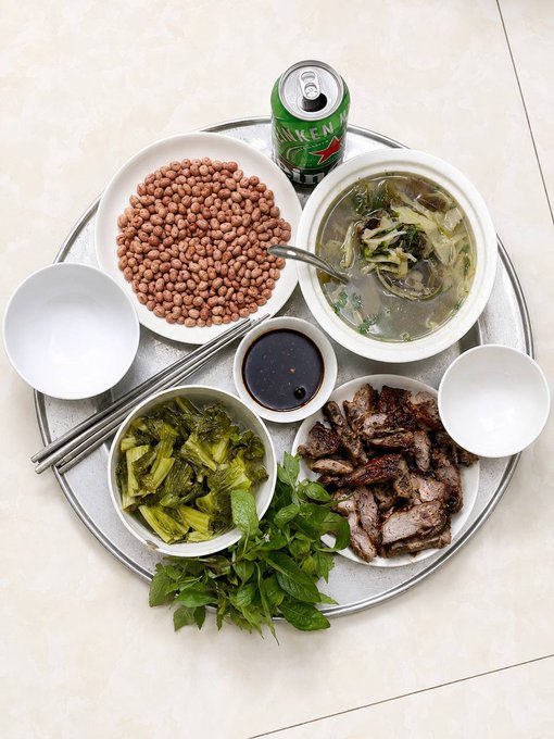 A Vietnamese meal spread on a round tray, including peanuts, grilled meat, pickled greens, a bowl of soup, dipping sauce, a Sprite can, and fresh herbs.