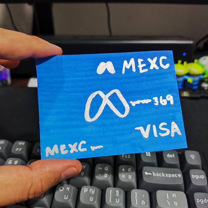 A person's hand holding a handmade blue card with white writing that reads 'MEXC', 'VISA', and '369' alongside an infinity symbol, held over a computer keyboard with small figurines visible in the background.