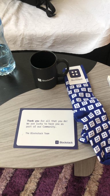 A photograph showing merchandise from 'Blockstack' on a round table, including a black mug with the Blockstack logo, a card reading 'Thank you for all that you do! We are lucky to have you as part of our community. The Blockstack Team', and a pair of blue Blockstack patterned socks.