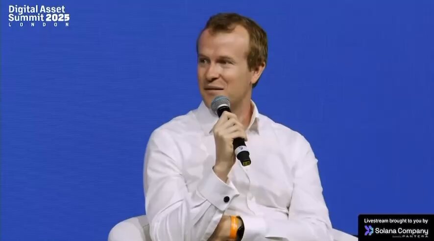 The image features a man sitting down with his arms crossed while holding a microphone. The background is blue, which could be an overhead projector screen or a large computer monitor displaying information related to cryptocurrency and blockchain technology. The caption of the image reads "Here to help #Crypto OGs Clouds (Data Centers) can offset security budgets and just use decentralized rails for transacting in the agentic economy and tokenizing their own products/ ai compute tokens. And @Google is doing it's homework."