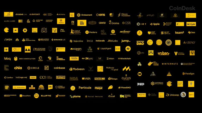 The image features a large, black and yellow poster displaying various logos of different companies and organizations. The poster includes logos for "Team1", "Team2", "Team3", "Team4", "Team5", "Team6", "Team7", "Team8", "Team9", "Team10", "Team11", "Team12", "Team13", "Team14", "Team15", and "Team16". The poster also includes logos for organizations such as "CoinDesk", "Team1", "Team2", "Team3", "Team4", "Team5", "Team6", "Team7", "Team8", "Team9", "Team10", "Team11", "Team12", and "Team13". The poster is set against a black background, making the logos stand out prominently.