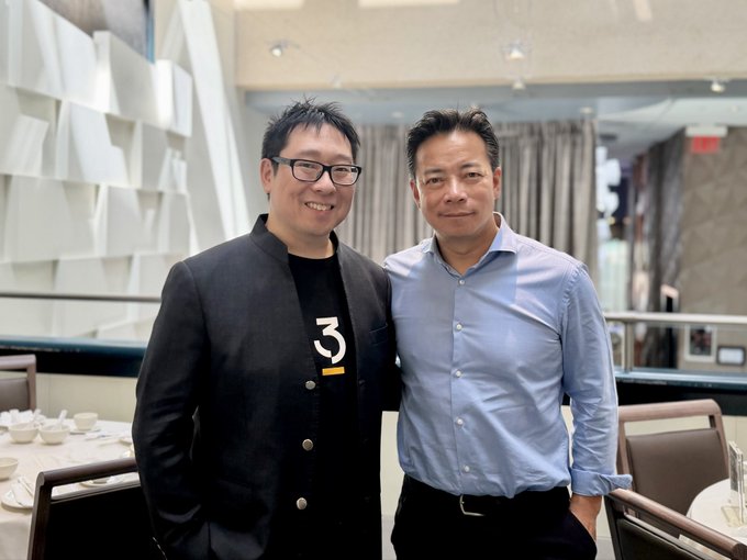 Two men posing together in what appears to be an upscale restaurant, with one wearing a black jacket over a t-shirt featuring a '3' logo and glasses, while the other wears a light blue button-down shirt.
