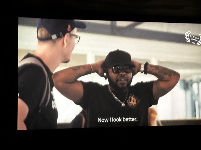 The image shows a man wearing glasses, a black hat, and a black shirt with the words "Now I look better" written above his head. The background of the image appears to be a television screen displaying an advertisement for Bitcoin Film Festival.