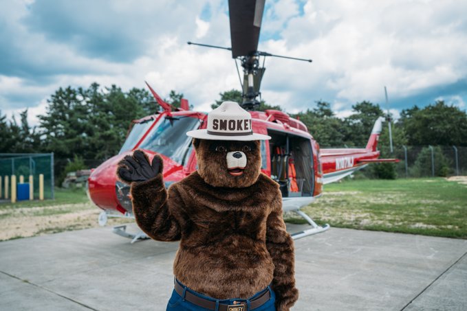 Smokey Bear mascot wearing a fire hat labeled 'SMOKE' standing in front of a red helicopter in an outdoor setting.