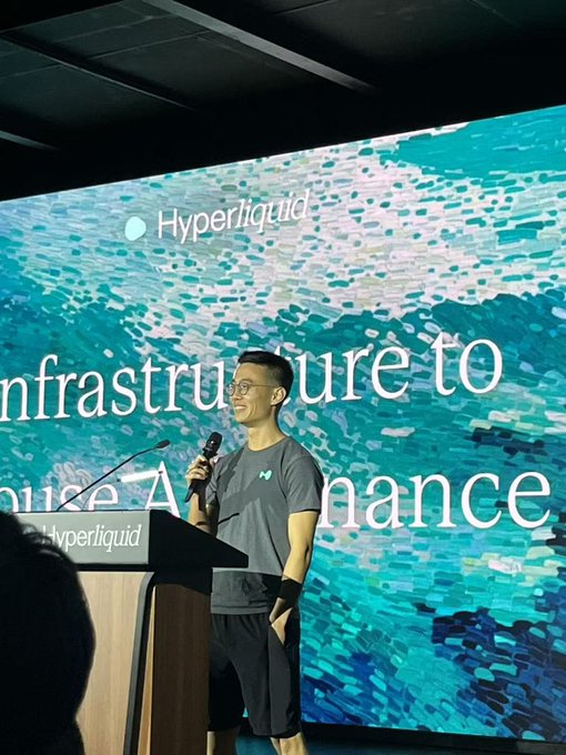 A man in glasses and a gray t-shirt speaks into a microphone at a podium during a Hyperliquid presentation, with a blue textured backdrop displaying 'Infrastructure to...nance'.