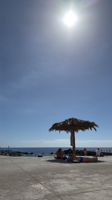 A sunny beach scene with a bright sun in a clear blue sky, featuring a thatched umbrella where people are relaxing on the concrete promenade by the ocean.