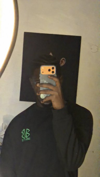 A person in a black "BBB" logo sweatshirt takes a mirror selfie with their face obscured by an iPhone in a peach-colored case, setting a casual "gm frens, get ready for today!" tone with their dark hoodie and grungy room backdrop.