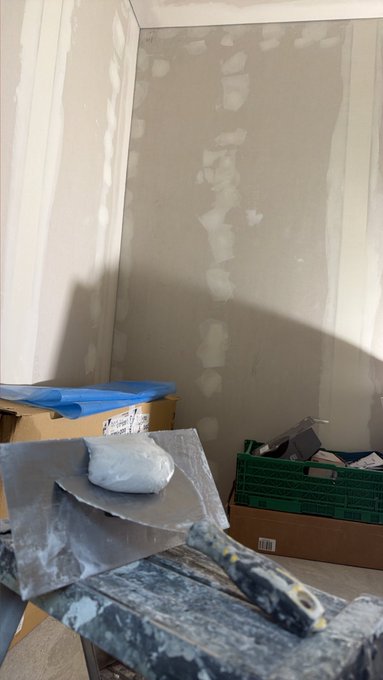 A home improvement scene shows unfinished drywall with joint compound patches, a putty knife resting on a drywall hawk, a green storage crate, and a cardboard box.