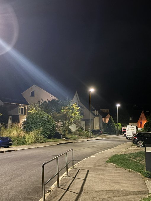 A nighttime street scene in a residential neighborhood, illuminated by streetlights, with houses, parked cars, and a metal barrier along the sidewalk.