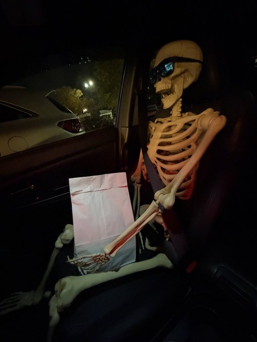 A skeleton wearing sunglasses sits in the passenger seat of a car at night, holding a white paper bag on its lap with its bony hands.