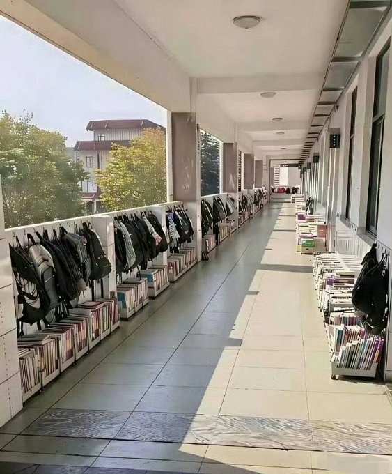 An outdoor hallway with backpacks hanging on the left wall and rows of books lined up along both sides, with trees and buildings visible in the background.