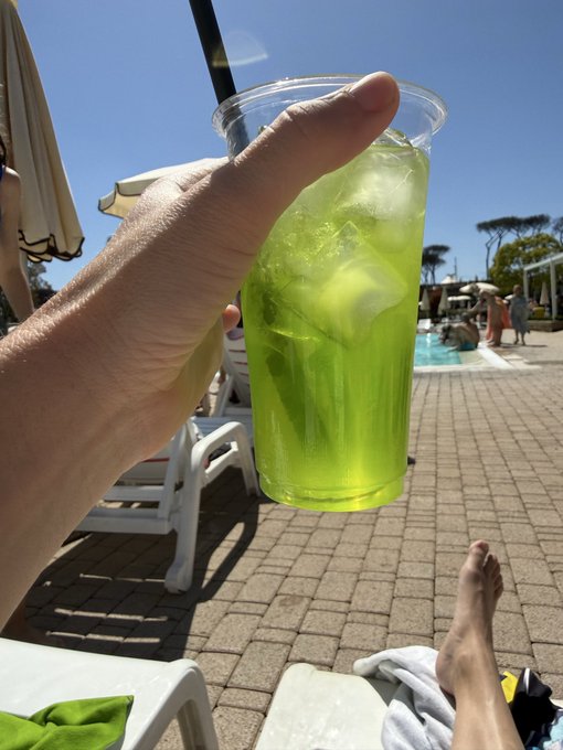 A hand reaches up with a vibrant green frozen cocktail in a plastic cup, set against a sunny poolside scene with lounge chairs and blue skies, perfectly capturing the relaxed vacation vibes of a late "gm."