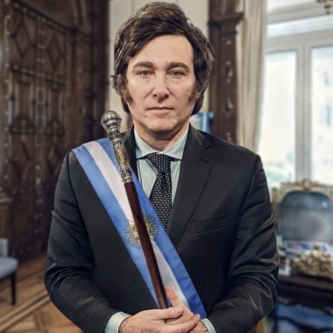 A man in a dark suit wearing the light blue and white presidential sash of Argentina stands in an ornate room, holding a ceremonial staff topped with a silver finial and featuring the Sun of May emblem. The image humorously connects to the post's theme by depicting Argentina's president—his country having embraced Bitcoin education on state TV—literally holding a "stick" symbolizing the nation's pivot toward BTC.
