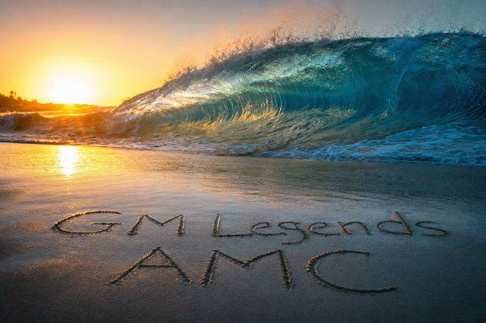 A scenic photograph of a large wave crashing on a beach at sunset, with "GM Legends AMC" written in the sand in the foreground.