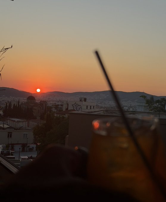 A sunset view over a city with mountains in the distance, featuring a cocktail glass with a straw silhouetted in the foreground.