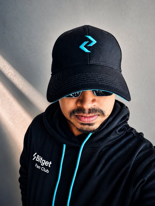 A person wearing a black 'Bitget Fan Club' hoodie and a black cap with a cyan logo, with the cap pulled down over sunglasses.