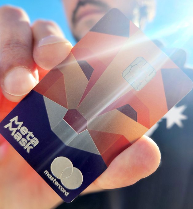 A hand holding a MetaMask-branded Mastercard credit card with a geometric orange and red design.