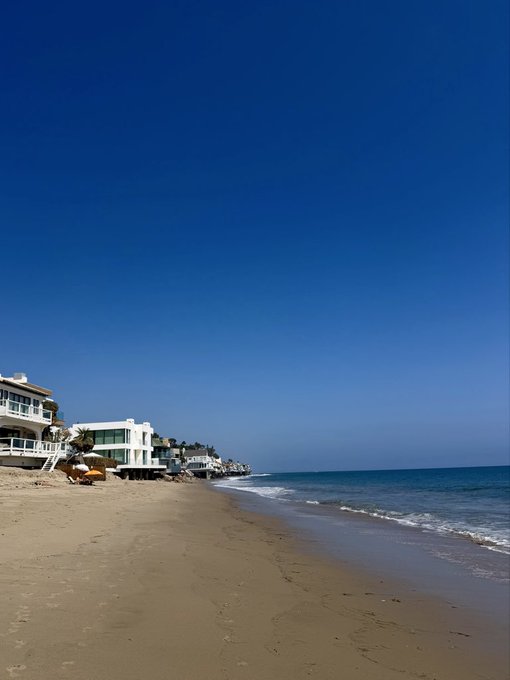 A serene beach scene with golden sand, gentle waves, and modern white houses lining the shore under a clear blue sky.