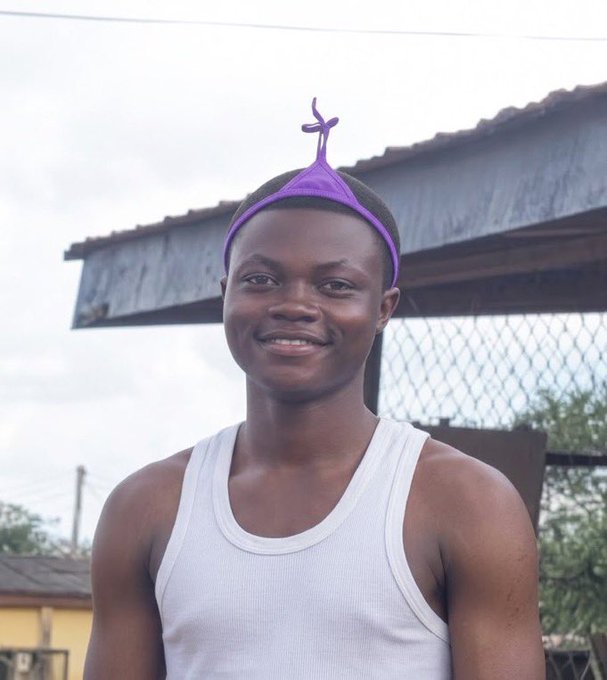 A smiling young man in a white tank top stands outdoors in front of a metal gate and building.