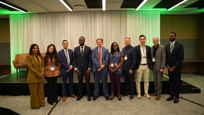 A diverse group of nine professionally dressed individuals pose together on a stage with a white curtain backdrop, representing representatives from ADAPT, the African Continental Free Trade Area (AfCFTA), the World Economic Forum (WEF), IOTA, and the Tony Blair Institute for Global Change (InstituteGC). The image visually captures the collaborative partnership highlighted in the post, showcasing the stakeholders working to advance public digital trade infrastructure across Africa.