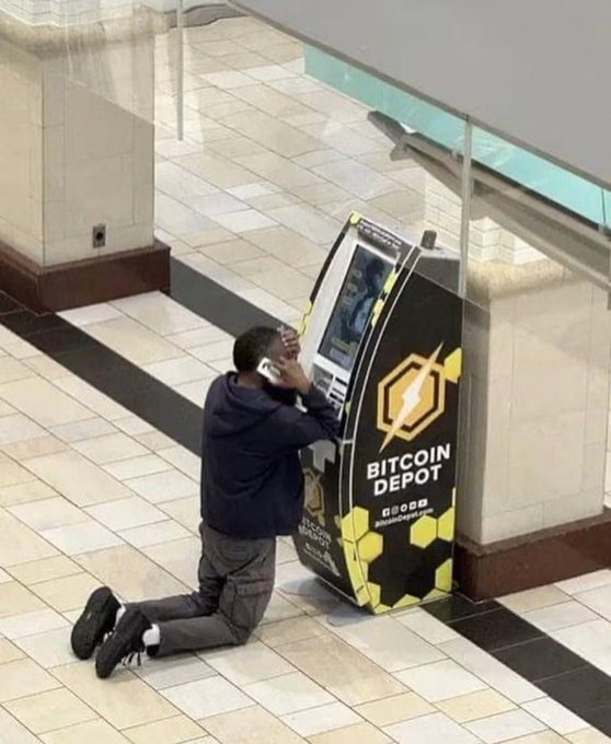 A man kneels on the floor of a shopping mall while talking on the phone in front of a "BITCOIN DEPOT" ATM, humorously illustrating the desperation of having missed out on the $ANSEM crypto pump.