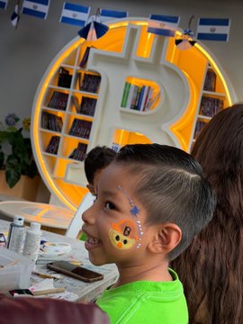 A young boy with face paint sits in front of a Bitcoin logo shelf and El Salvador flags.