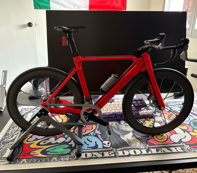 A red LEGO Technic model bicycle with black wheels displayed on a colorful "One Dollar" mousepad in front of a monitor, keyboard, and Italian flag.