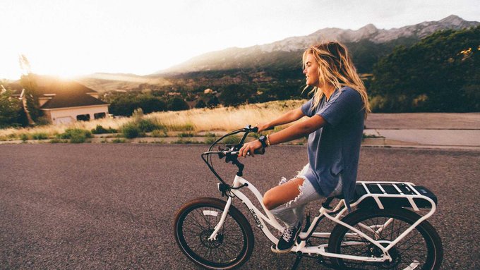 A blonde woman wearing a blue t-shirt and denim shorts smiles while riding a white electric cargo bike along a paved road, with sunlit mountains and a house visible in the background.