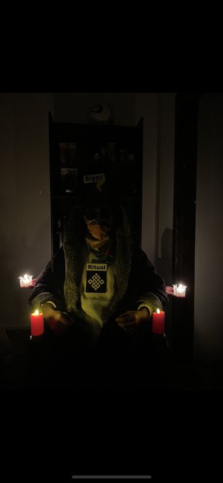 A dimly lit photo of a person seated in a chair holding lit candles, wearing a dark hooded jacket and a shirt with the word 'Ritual' printed on it.