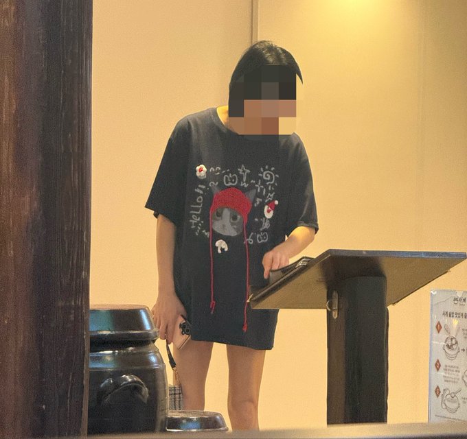 A person wearing a black Hello Kitty t-shirt dress stands at a wooden podium, with their face blurred for privacy.