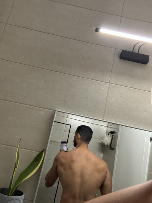 A shirtless man takes a mirror selfie in a bathroom, holding his phone while a snake plant sits in the corner, accompanying a social media post asking for a quote response.