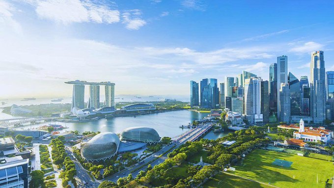 An aerial view of Singapore's Marina Bay skyline featuring the iconic Marina Bay Sands hotel, ArtScience Museum, Esplanade, and Central Business District skyscrapers under a bright blue sky.