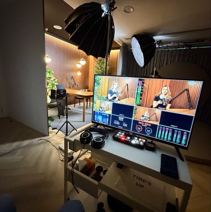 A professional streaming/podcast setup featuring a monitor displaying a multi-camera view of a woman at a microphone, accompanied by studio lighting, a keyboard mixer, and headphones on a white desk with 'TIMES UP' written below.