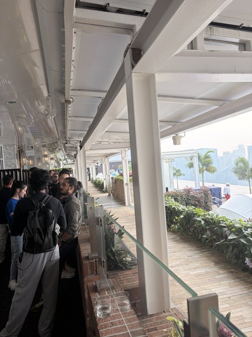 Several people wait in line at an outdoor covered venue with a glass railing, overlooking a waterfront area with plants.