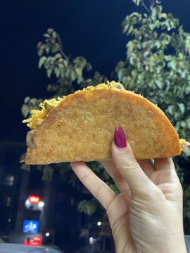 A hand with red nail polish holds up a halved, crispy fried taco or sandwich against a nighttime background with a tree and blurred street lights.