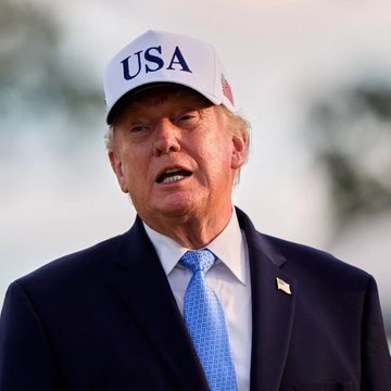 A man wearing a "USA" cap and dark blue suit with a light blue tie speaks outdoors, his face partially in shadow against a hazy sky. The caption relates to news that the man pictured, Donald Trump, is considering former Director of National Intelligence Jay Clayton for an AI Czar role.