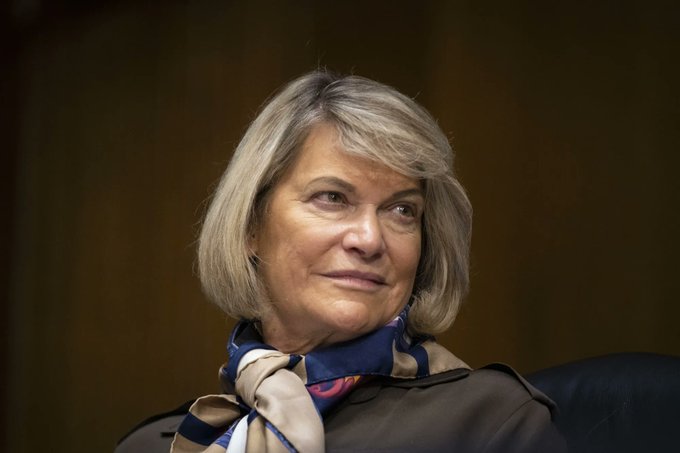 A woman with shoulder-length gray hair wearing a brown blazer and a blue, beige, and pink patterned scarf looks off to the side with a slight smile, appearing composed in what seems to be an official setting. Her polished appearance suggests she may be a public figure or politician, fitting the context of the accompanying post about Senator Cynthia Lummis criticizing Democrats after the CLARITY vote.