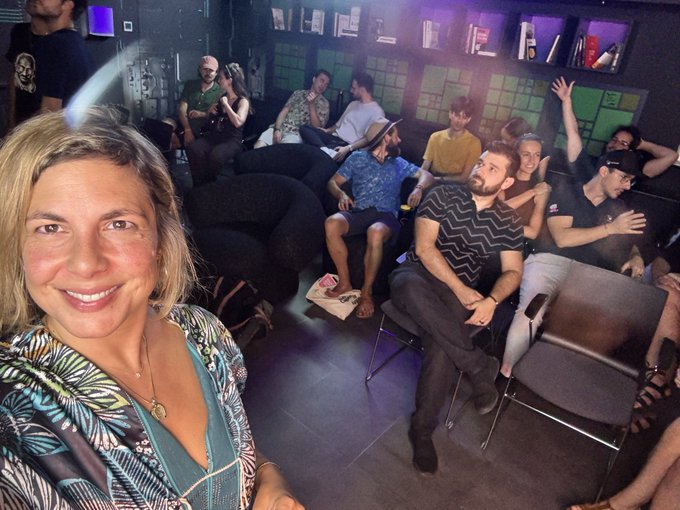 A woman takes a selfie in the foreground, smiling, while a group of attendees sit on couches and chairs behind her in a dimly lit lounge, gathered for a screening. The setting reflects the post's description of a peer-to-peer viewing event for the Julian Assange documentary "Six Billion Dollar Man," hosted by a freedom and bitcoin community gathering.
