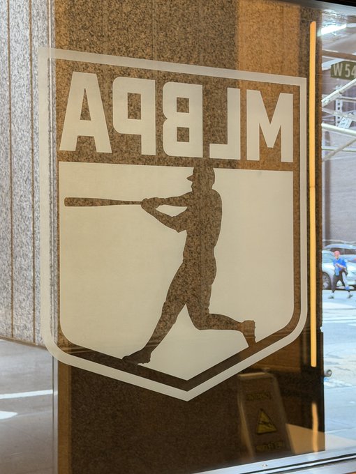 A logo etched into a marble wall displays "A98JM" above a silhouette of a baseball batter swinging a bat, appearing to be the MLB shield emblem. The image connects to the news by visually representing the MLB's entry into the NFT marketplace on OpenSea, with "A98JM" likely serving as a reference to the Solona wallet address used for the minting.